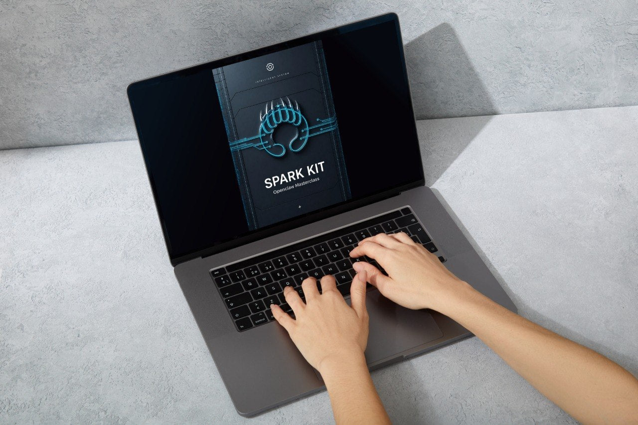 A dark gray laptop sits open on a light gray tabletop against a textured gray wall. The screen displays a digital cover titled 'SPARK kIT' with orange graphic accents.